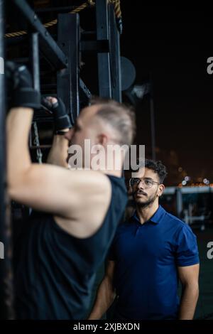 A man is doing pull-ups on a rig, while his trainer watches. Stock Photo