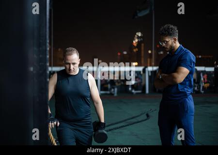 A man wearing a black tank top and black shorts lifts weights while a man wearing a blue shirt watches. Stock Photo