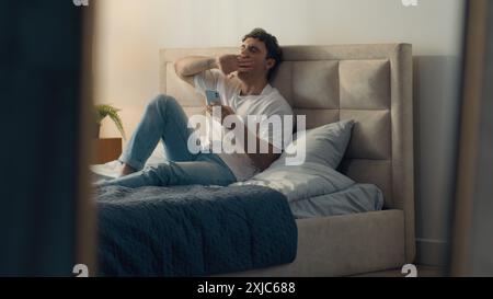 Man sitting on bed bedroom using mobile phone smartphone telephone yawning lazy male guy network browsing web electronic gadget home weekend bedding Stock Photo