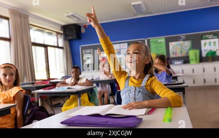 Caucasian schoolgirl raising hand while studying with diverse children in classroom. Unaltered, education, student, german, school, childhood, learnin Stock Photo