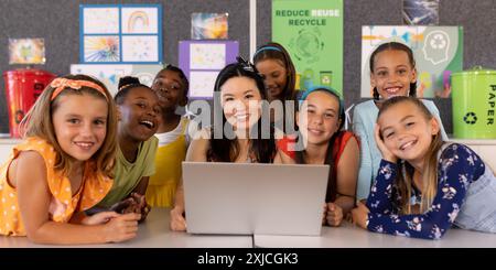 Portrait of smiling diverse female teacher and schoolgirls with laptop on desk in classroom Stock Photo