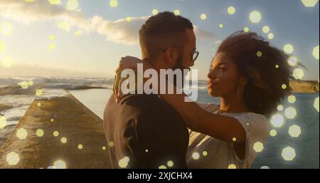 Image of spots of light over happy diverse couple embracing by sea Stock Photo
