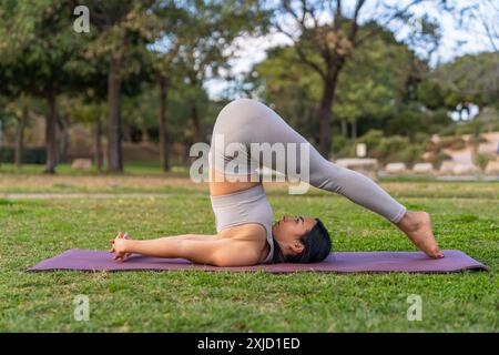 A brunette woman dressed in sportswear doing halasana yoga posture or plow pose in the park. Stock Photo