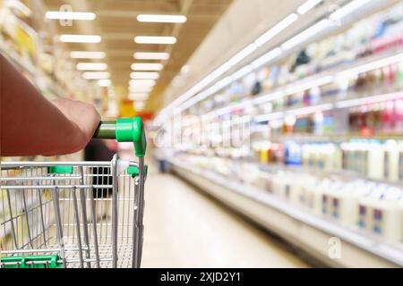 woman hand hold supermarket shopping cart with abstract blur refrigerator shelves with fresh milk bottles and dairy products in grocery store Stock Photo