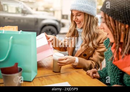 Excited young women looking at shopping bags content. Diverse friends sharing moment of joy during coffee break after successful shopping trip - Frien Stock Photo