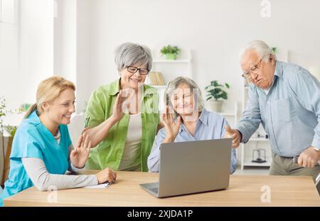Senior people sitting in nursing home with nurse, waving hand having online video call using laptop Stock Photo