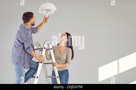 Young man standing on the ladder changing light bulb in the room with his smiling wife. Stock Photo