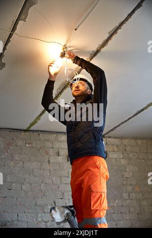 Electrician wearing a white safety helmet and protective glasses, standing on the ladder focused on wiring a light bulb fixture on the ceiling. Stock Photo