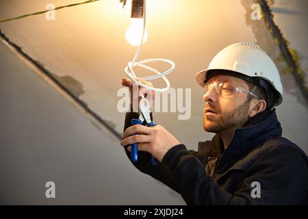 Portrait of male electrician, in the process of wiring a temporary light bulb fixture on the ceiling and cutting white cable with wire cutters. Stock Photo
