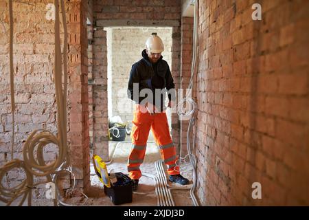 Electrician using wire cutters during installation of electrical wiring, on building under renovation, with exposed brick walls and unfinished surface Stock Photo