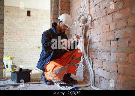 Construction worker installing electrical wiring, using wire cutters, inside building under construction with exposed brick walls and unfinished surfa Stock Photo