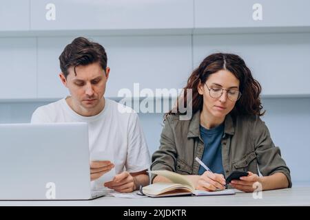 Couple managing their finances at home, using a laptop and smartphone, reviewing receipts, and taking notes in a bright kitchen. Husband and wife plan the family budget and spending money.  Stock Photo