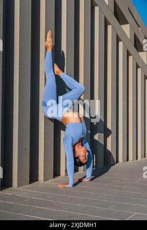A vertical shot of a woman in blue active-wear performing a handstand against a modern concrete wall on a sunny day Stock Photo