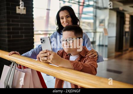 A mother and her son with Down syndrome are smiling and using a phone while shopping in a mall. Stock Photo