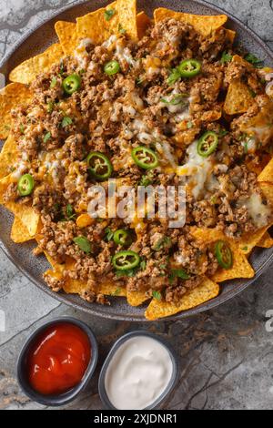 Mexican nacho corn tortilla chips with meat, cheddar cheese and jalapeno  pepper served with sauces closeup on the plate on the table. Vertical top vi Stock Photo