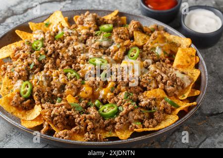 Corn nachos with lots of stewed minced meat, cheddar cheese and jalapeno  pepper served with sauces closeup on the plate on the table. Horizontal Stock Photo