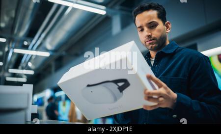 Handsome Young Man Shopping for a Virtual Reality Computer in a Home Electronics Store. Multiethnic Customer Comparing Different Headset Technology Features, Sale Price and VR Quality of the Device Stock Photo