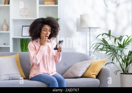 Woman with surprised expression sitting on sofa holding smartphone. Bright modern living room with plants and shelves in the background. Concepts of unexpected news, surprise, shock, and technology. Stock Photo