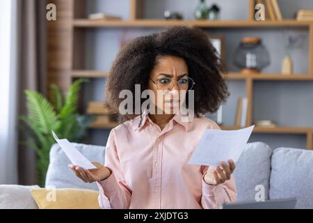Young woman with glasses and curly hair looks confused as she reviews financial documents. Sitting on couch in a cozy living room, she appears frustrated with bills or paperwork. Stock Photo