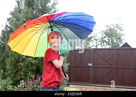 child with a rainbow umbrella stands under a summer shower. On a cloudy day, a 7-year-old boy stands under a multi-colored umbrella, with rain falling Stock Photo