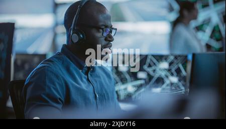 Black Man Effectively Multitasking with Supporting Clients on a Phone Call and Computer Work. Handsome Surveillance Specialist in a Modern Monitoring Room with Displays and Computers Stock Photo