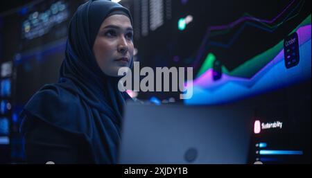 Muslim Female Data Center IT Engineer Standing in a Room with Statistics and Charts Settings on a Digital Screen. Cloud Computing Expert Uses Laptop for Servicing the System in Cyber Security Facility Stock Photo