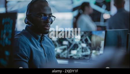 African Specialist in a Dark High-Tech Monitoring Room, Concentrated on a Call and Computer Work. Black Technical Support Manager Using Headphones with Built-In Mic for Communication with Client Stock Photo