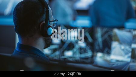 Technical Customer Support Specialist is Talking on a Headset while Working on a Computer in a Call Center Control Room Filled Display Screens and Data Servers. Footage from the Back Stock Photo