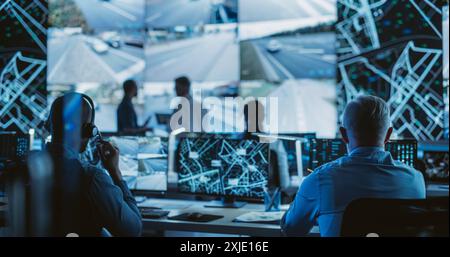 Dark Room with Multiethnic Specialists Working on Desktop Computers. Big Digital Screen Shows Surveillance Satellite Data, City Map, and CCTV Traffic Footage Reports Stock Photo