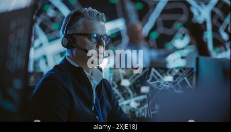 Portrait of a Middle Aged Logistics Officer Wearing a Headset, Working in a Central Office Hub for Delivery Operations, Control and Monitoring for Managing International Fleet of Drivers Stock Photo