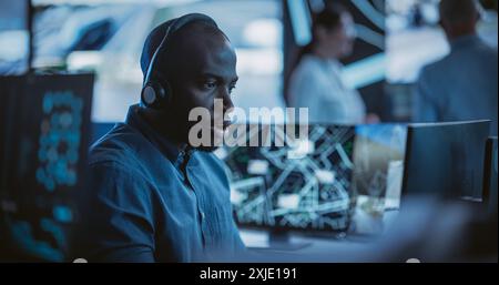 Handsome Surveillance Specialist in a Modern Monitoring Room with Displays and Computers. Black Man Effectively Multitasking with Supporting Clients on a Phone Call and Computer Work Stock Photo