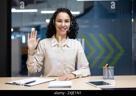 Businesswoman wearing headset sitting at office desk waving with cheerful expression, engaging in virtual meeting. Modern workspace in background, focus on communication and technology. Stock Photo