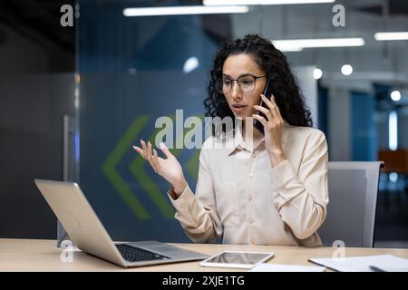 Businesswoman in office on phone call working on laptop. Professional woman multitasking, with digital tablet and paperwork on desk. Modern office environment with contemporary design. Stock Photo