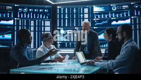 Team Of Data Scientists and Software Developers Discussing Reports in Monitoring Room with Analytics Feed on Big Digital Screen. Senior Man Leading Artificial Intelligence Company Board Meeting Stock Photo