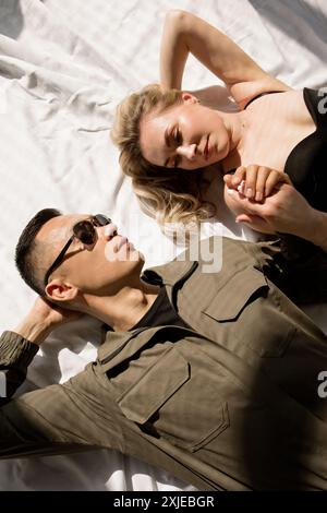 A man in a uniform and sunglasses and a woman lie on a bed holding hands in the sun Stock Photo