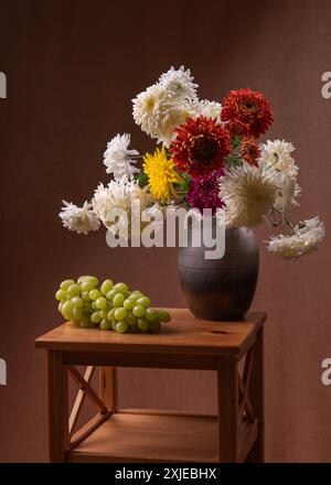 large bouquet of white, yellow, red, autumn flowers. Many asters in a gray vase on a wooden table, next to it lies a large bunch of green grapes. Oran Stock Photo