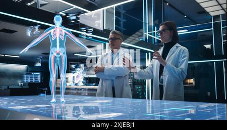 Two Bioengineers Working With Computer-Powered VFX Hologram Of Human Body And Organs In Futuristic Lab. Man And Woman Researching Blood Flow, Developing Innovative Technological Healthcare Solutions. Stock Photo