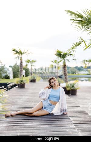 A woman sits on a wooden dock, wearing a white kimono and blue denim shorts. She is looking at the camera and has one hand on her hip. Palm trees and Stock Photo