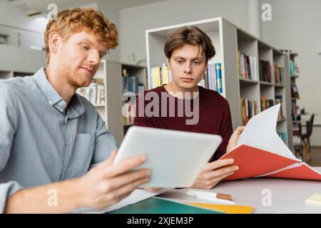 Two students engage with a tablet while seated at a table in a library. Stock Photo