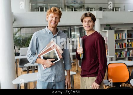 Two young male students stand side by side, each holding a book in a library. Stock Photo