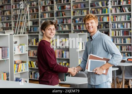 Two young men shake hands in a quiet library, surrounded by books and studying materials. Stock Photo