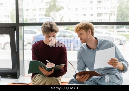 Two male students seated on a bench, engaged in studying papers together. Stock Photo