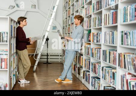 Two young male students delve into books while standing near a library ladder. Stock Photo