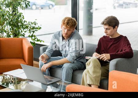 Two men engrossed in work, sitting on couches with laptops. Stock Photo