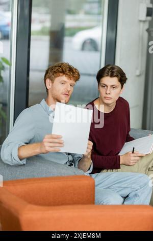 Two male students deeply engaged in studying papers on couch. Stock Photo