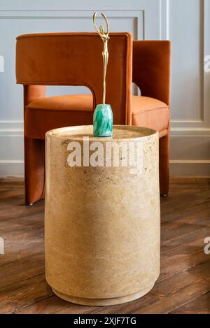 Marble side table with a gold sculpture on top and orange modern chair in the background. The table is a light beige color and the sculpture has a green base Stock Photo