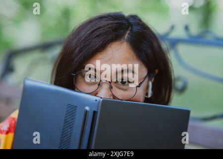 Laughing woman combines study with fun, using closed laptop to shade herself. Asian woman with glasses having fun with her laptop outdoors. Stock Photo
