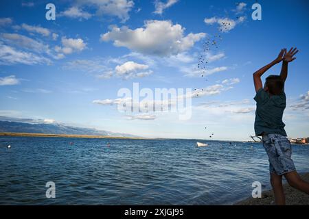 Young boy throwing stones into a calm blue sea on a summer day with white clouds in the sky. Stock Photo