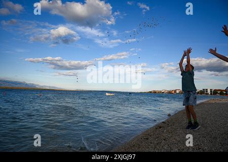 Young boy throwing stones into a calm blue sea on a summer day with white clouds in the sky. Stock Photo