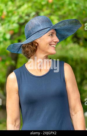 elderly beautiful woman 50-55 years old in a blue lady's hat and dress laughing in a summer garden Stock Photo